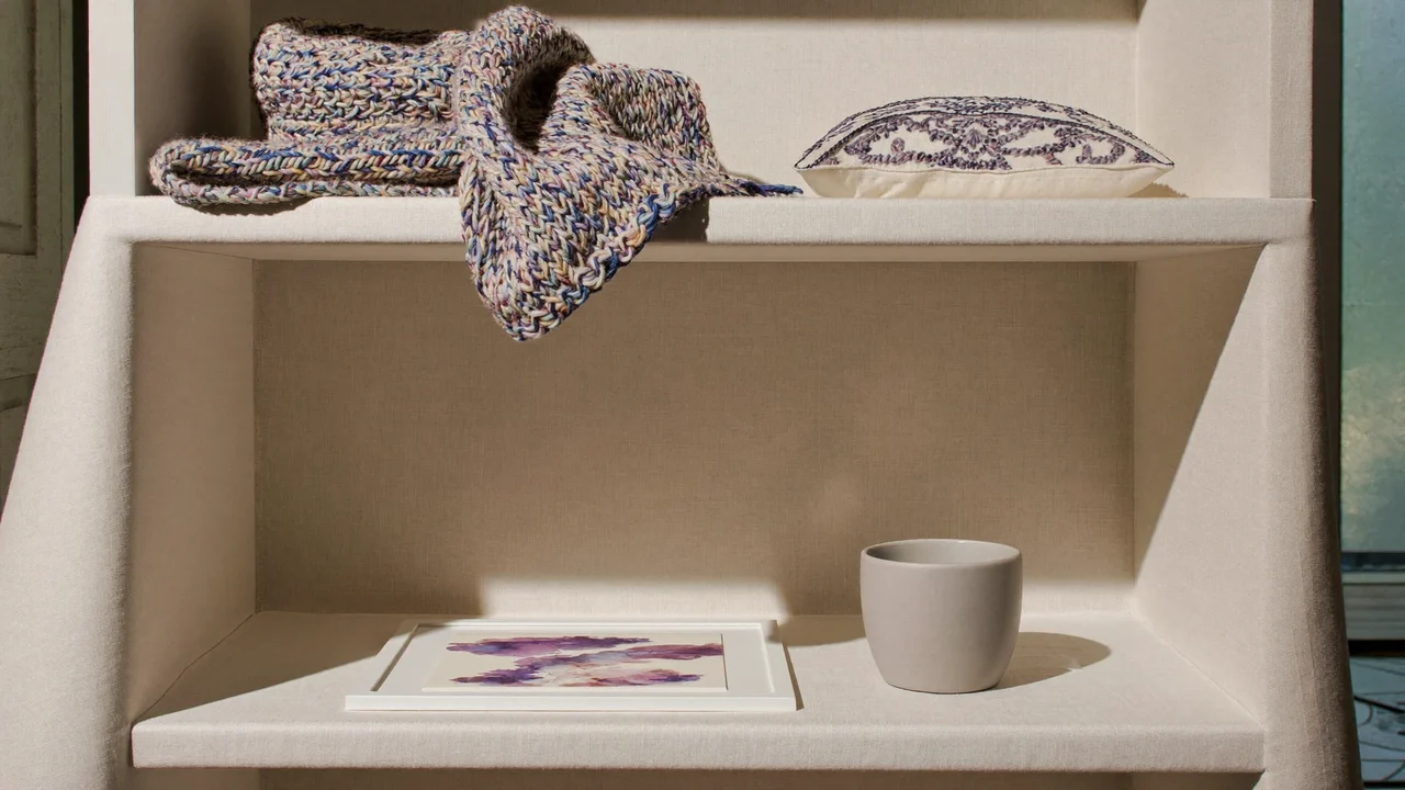 Scialle, cuscino ricamato, tazza in ceramica e acquerello esposti su una mensola in lino.